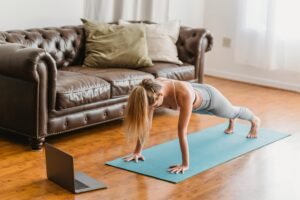 Side view full body of sportive female in activewear practicing kumbhakasana asana on mat near laptop during online lesson in living room near couch